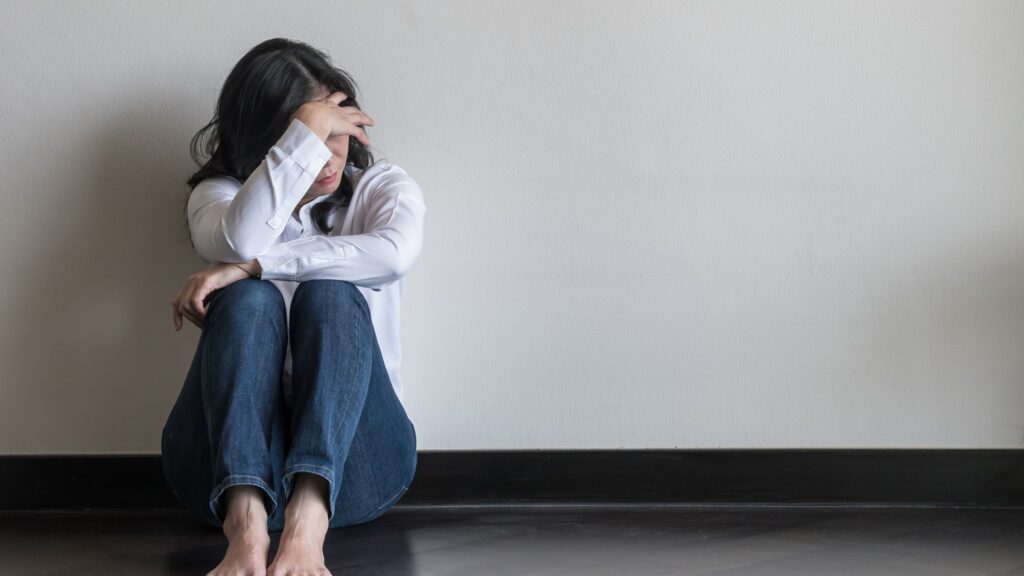 This image shows a woman with her head in her hands, sat with her back against a white wall. - ADHD rejection sensitivity dysphoria