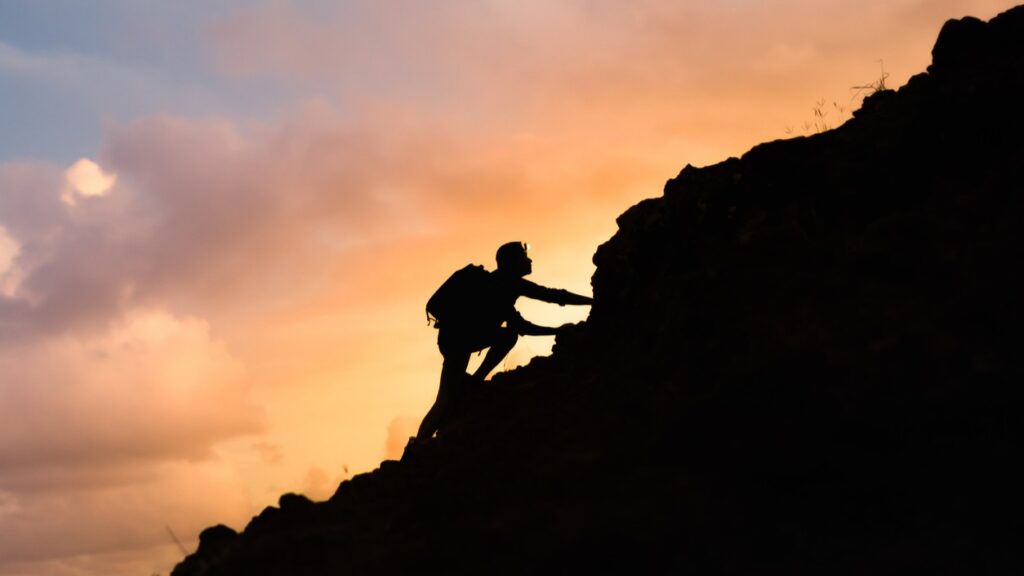 This image shows a silhouette of a person climbing up a hill with a sunset in the background. - On the Harder Days Gentle Reminders for Your Mind and Body - self compassion reminders