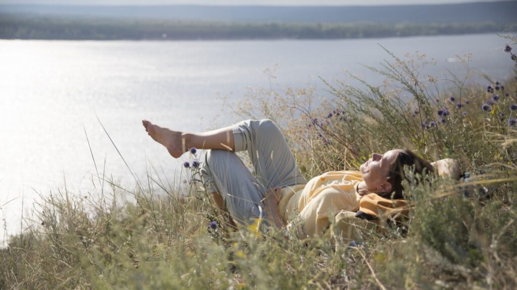 This image shows a woman lying on a grassy bank overlooking the sea. - Why is it so hard to rest